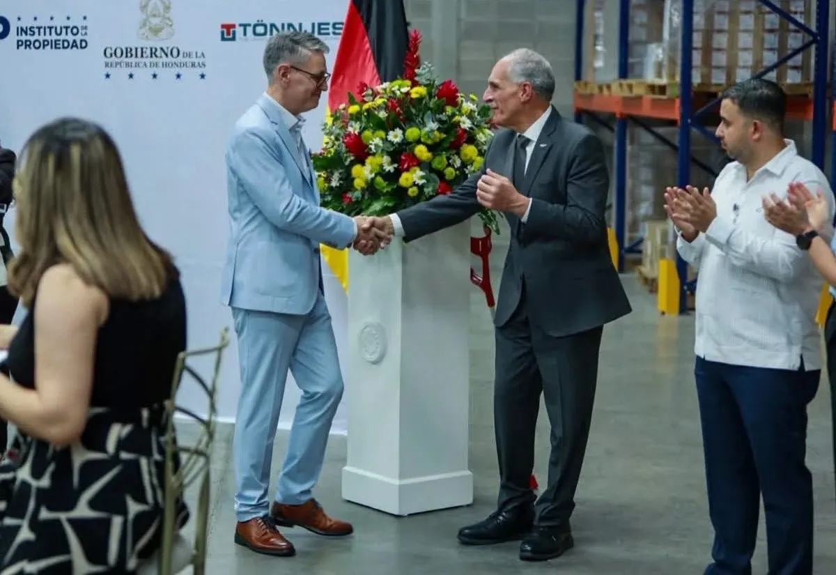 Dos representantes se dan la mano junto a un arreglo floral durante el acto de arranque de la producción de placas, entre aplausos de los asistentes