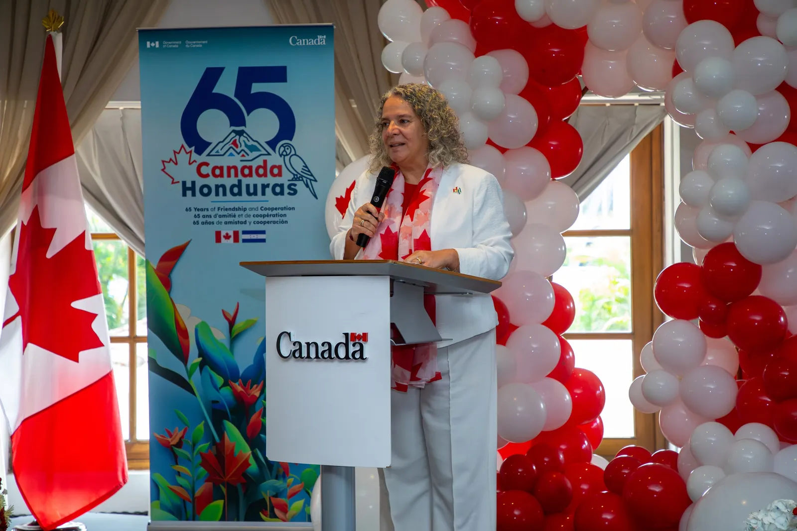 Otra oradora se dirige al público desde el atril, entre la bandera de Canadá y el cartel del 65 aniversario