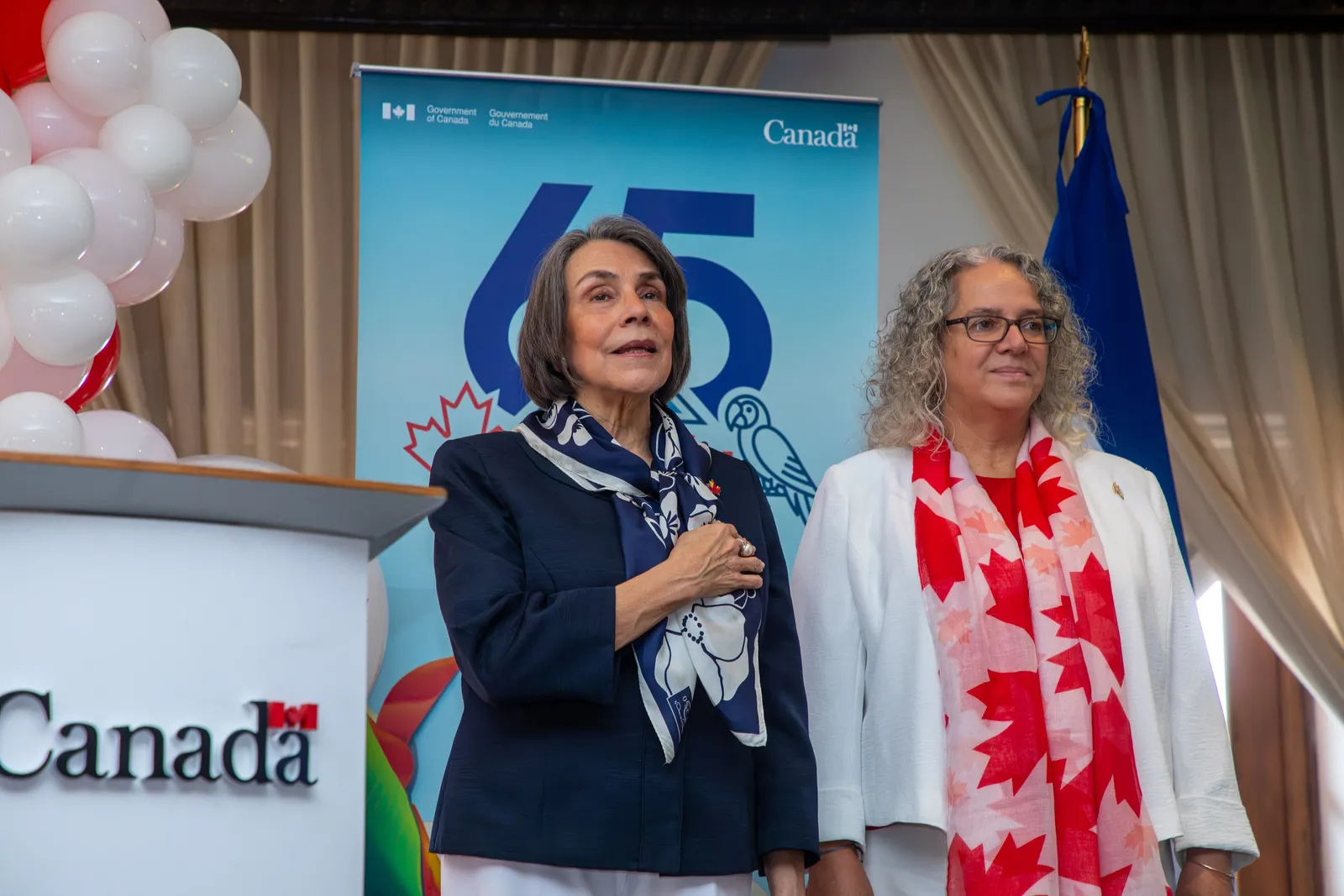Dos participantes en el escenario, una con la mano en el pecho, frente al cartel del 65 aniversario de relaciones entre Canadá y Honduras