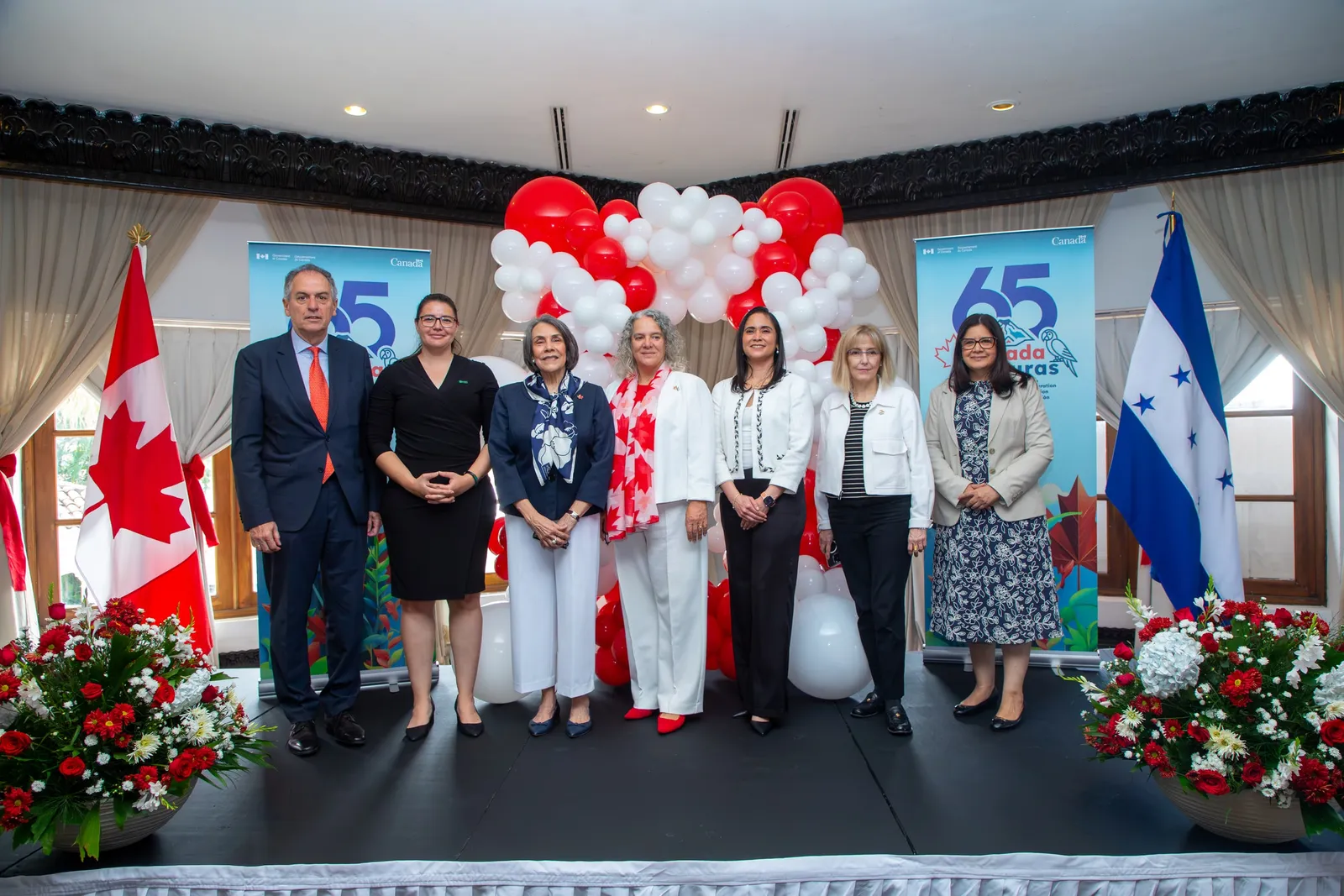 Foto de grupo en el escenario, entre las banderas de Canadá y Honduras, durante la celebración del 65 aniversario